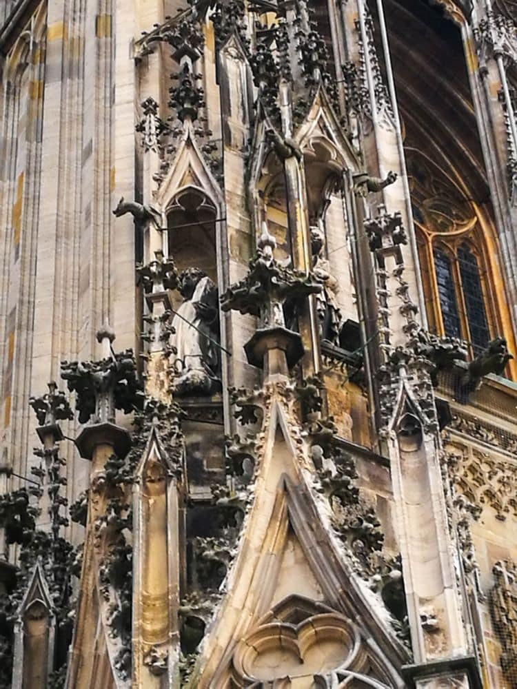 Detaillierte Nahaufnahme der gotischen Fassade des Ulmer Münsters mit kunstvollen Steinmetzarbeiten, Spitzbögen, zahlreichen Statuen und dekorativen Türmen. Im Hintergrund ist ein Teil eines Glasfensters zu sehen.
