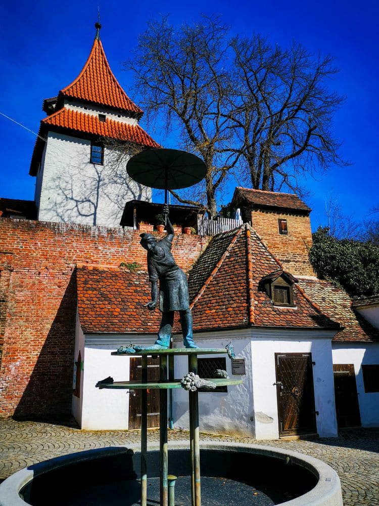 Die Bronzestatue eines Mannes, der einen Regenschirm hält, steht auf einem Sockel in einem kleinen runden Brunnen in Ulm, umgeben von alten Backsteinbauten mit roten Ziegeldächern und einem hohen, blattlosen Baum unter einem strahlend blauen Himmel.