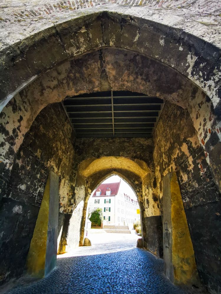 Blick aus einem alten, steinernen Torbogen mit verwitterten Mauern in Ulm auf eine sonnenbeschienene Straße und weiße Gebäude mit roten Dächern im Hintergrund.