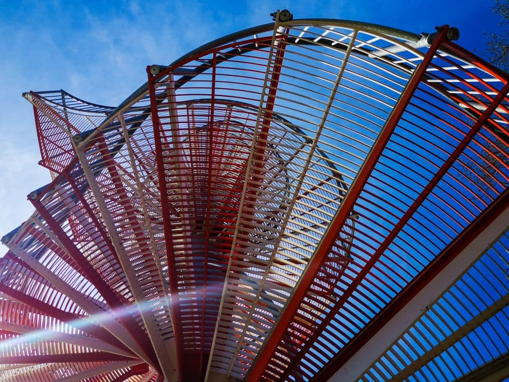 Gebogene Metallstruktur mit roten und silbernen Stäben, die ein spiralförmiges Muster bilden, von unten gesehen vor einem strahlend blauen Ulmer Himmel. Das Sonnenlicht erzeugt ein Streulicht in der unteren linken Ecke des Bildes.