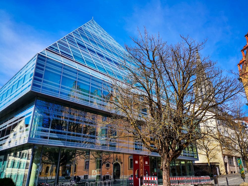 Ein modernes Glaspyramidengebäude mit spiegelnden Fenstern steht neben einem blattlosen Baum und historischer Ulmer Architektur unter strahlend blauem Himmel.