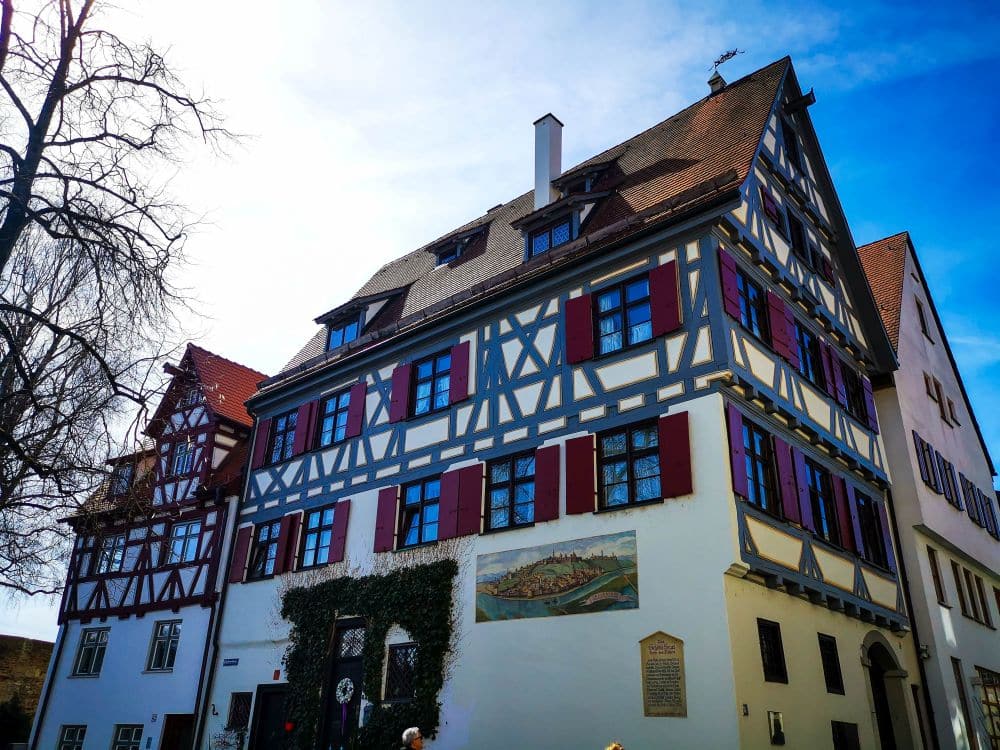 Ein traditionelles Fachwerkhaus in Ulm mit roten Fensterläden und einer Wandmalerei auf der weißen Fassade steht unter blauem Himmel. Auf der linken Seite des Bildes sind blattlose Äste zu sehen.