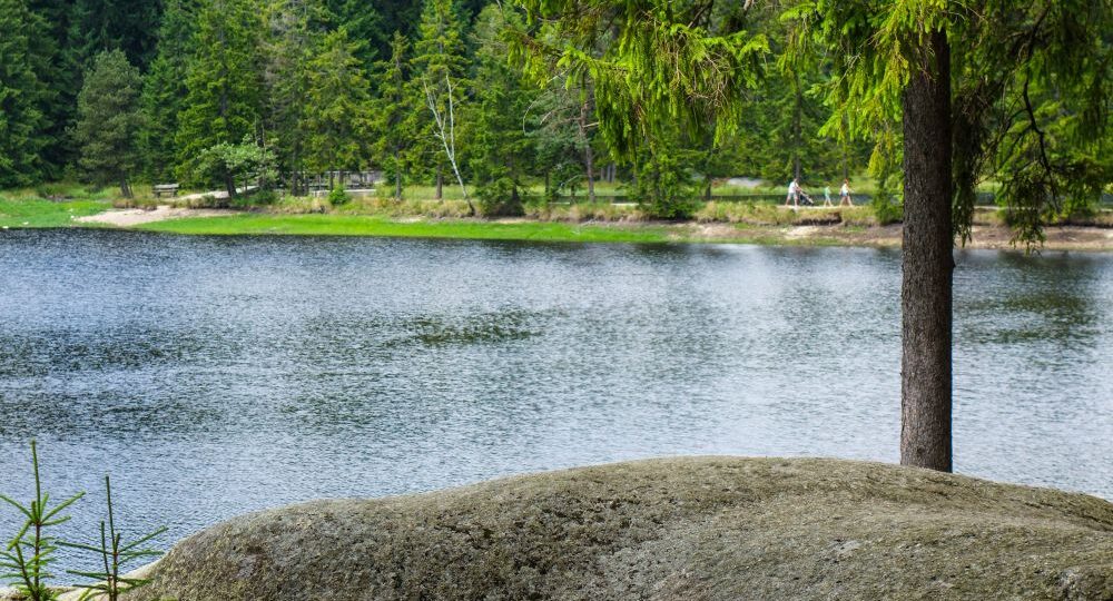 Ein großer Felsen liegt neben dem ruhigen Fichtelsee, der von dichten grünen Bäumen gesäumt wird. Im Hintergrund spazieren ein paar Menschen auf einem Weg in der Nähe des Ufers.