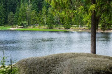 Ein großer Felsen liegt neben dem ruhigen Fichtelsee, der von dichten grünen Bäumen gesäumt wird. Im Hintergrund spazieren ein paar Menschen auf einem Weg in der Nähe des Ufers.