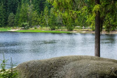 Ein großer Felsen liegt neben dem ruhigen Fichtelsee, der von dichten grünen Bäumen gesäumt wird. Im Hintergrund spazieren ein paar Menschen auf einem Weg in der Nähe des Ufers.