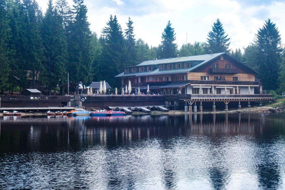 Eine große Holzhütte liegt am ruhigen Fichtelsee, umgeben von Kiefern. Am Ufer liegen bunte Paddelboote, und eine Außenterrasse mit Tischen bietet einen Blick auf das Wasser, in dem sich das Gebäude und die Bäume spiegeln.