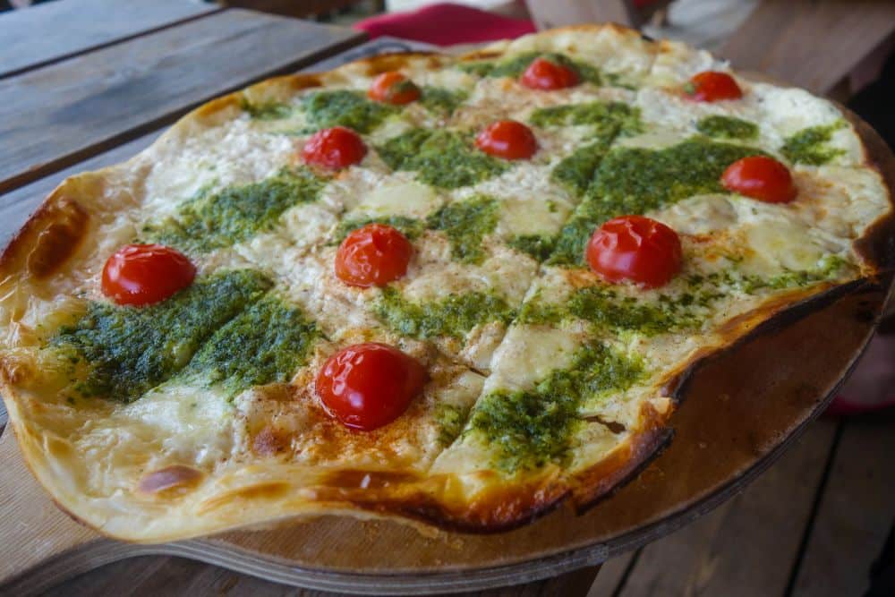 Eine Pizza mit dünner Kruste, belegt mit geschmolzenem Käse, grüner Pestosauce und ganzen Kirschtomaten, serviert auf einem Holzbrett - perfekt, um sie am malerischen Fichtelsee zu genießen.