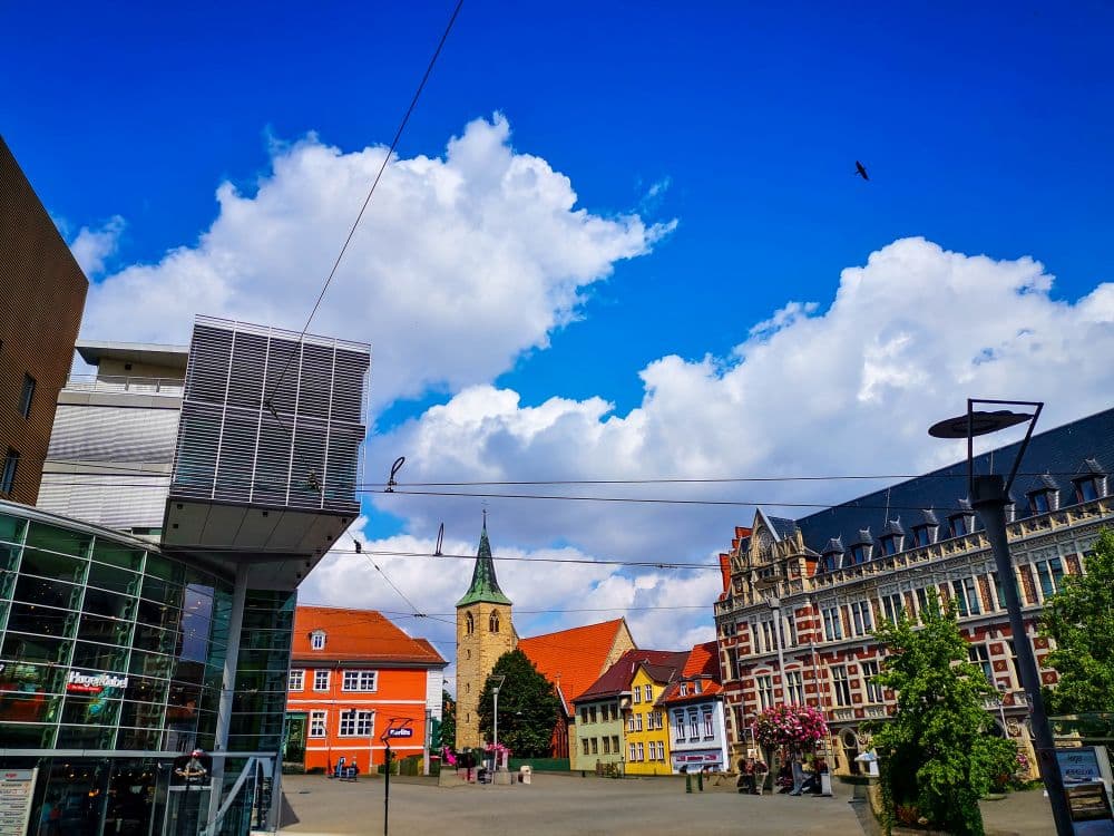 Ein lebendiger Stadtplatz in Erfurt mit farbenfrohen historischen Gebäuden, einer Kirche mit einem hohen Kirchturm, modernen Glasbauten, Straßenlaternen und einem strahlend blauen Himmel voller flauschiger Wolken. Ein Vogel fliegt hoch in den Himmel.