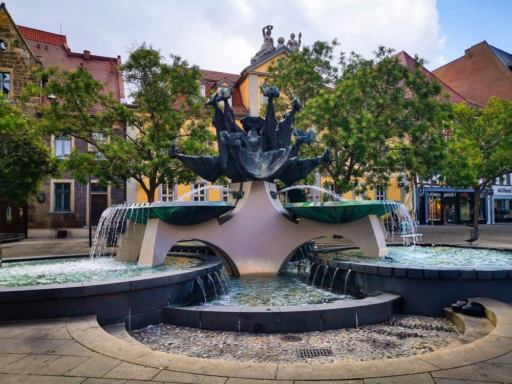 Auf einem Erfurter Stadtplatz, umgeben von Bäumen und historischen Gebäuden mit roten Ziegeldächern, steht an einem teilweise bewölkten Tag ein Zierbrunnen mit skulptierten Figuren und mehreren Wasserströmen.