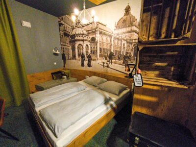 Ein gemütliches Hotelzimmer mit einem Doppelbett, Holzmöbeln, einem Kronleuchter und einem großen Wandgemälde, das die Jugendstilarchitektur Wiesbadens und das historische Stadtleben darstellt - perfekt für die Erkundung der berühmten Thermen der Stadt.