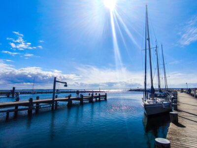 Die Sonne strahlt über dem ruhigen Hafen von Poel, in dem Segelboote an einem hölzernen Pier vor Anker liegen, der Himmel klar und blau ist, die Wolken verstreut sind und die Spiegelungen auf dem Wasser schimmern.