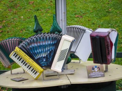 Mehrere bunte Klingenthaler Akkordeons sind in einem Halbkreis auf einem runden Tisch im Freien angeordnet, mit grünem Gras im Hintergrund. Zwischen den Instrumenten ist ein kleines Schild mit deutscher Schrift angebracht.