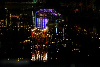 Während der Lichterserenade treiben mit bunten Lichtern geschmückte Boote auf einem nächtlichen Fluss, umgeben von zahlreichen kleinen, leuchtenden Laternen, die sich auf dem Wasser spiegeln und eine festliche und heitere Atmosphäre schaffen.