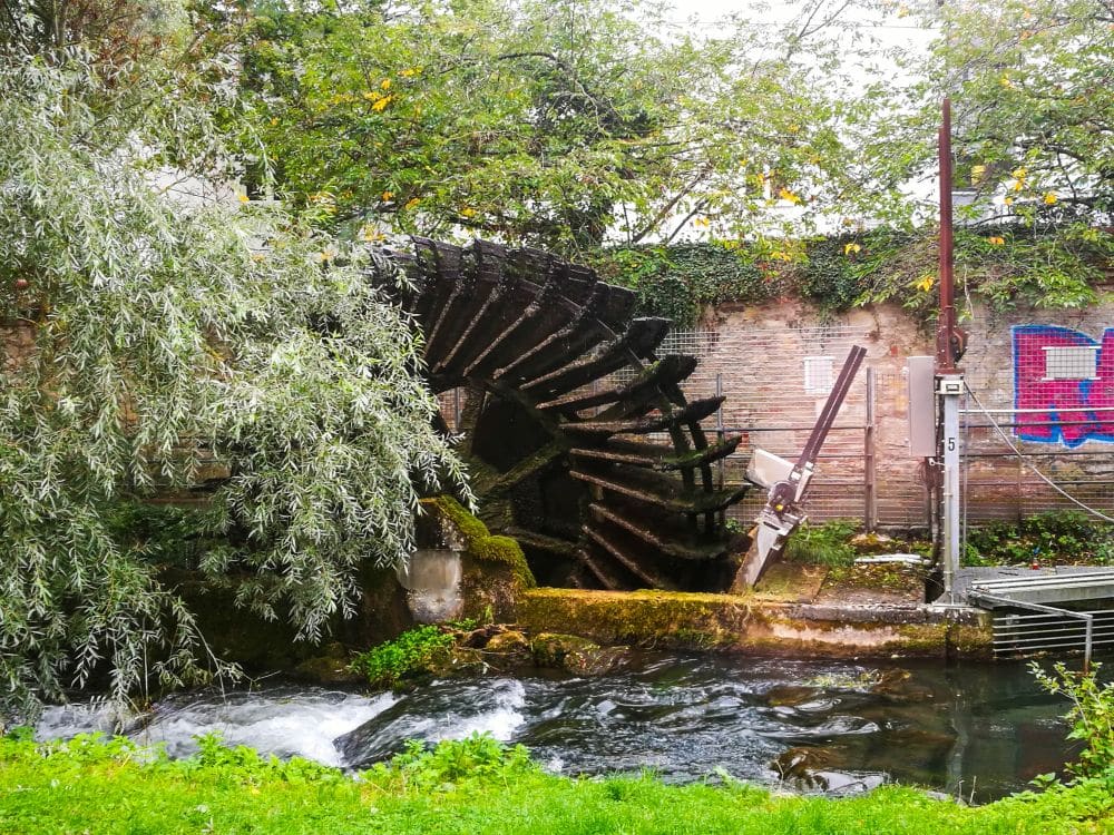 Ein großes hölzernes, moosbewachsenes Wasserrad steht neben einem fließenden Bach, umgeben von grünem Gras, Bäumen und einer Backsteinmauer mit bunten Graffiti - ein malerischer Ort, der oft auf der Ulm Card Tour zu sehen ist.