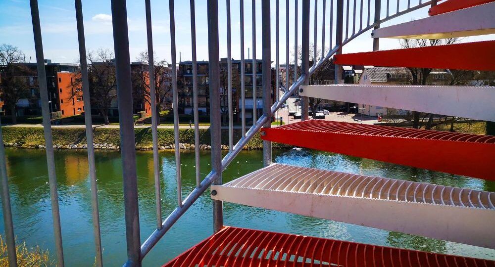 Im Vordergrund ist eine rot-weiße Metalltreppe zu sehen, die auf einen Fluss mit ruhigem Wasser blickt. Auf der anderen Seite des Flusses stehen Ulmer Sehenswürdigkeiten wie moderne Wohnhäuser und blattlose Bäume unter einem blauen Himmel.