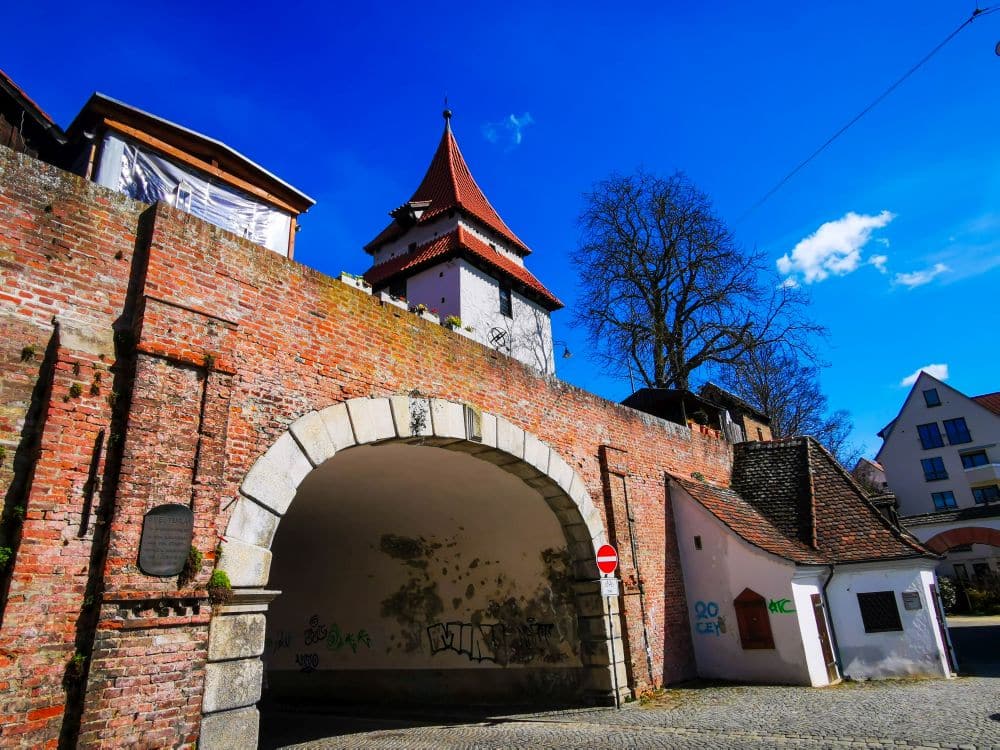 Ein historischer Backsteinbogen mit einem Turm darüber steht unter einem strahlend blauen Himmel. An der unteren Wand sind einige Graffiti zu sehen, und eine Straße mit Kopfsteinpflaster - die Sie mit Ihrer Ulm Card befahren können - führt durch den Bogen an alten Gebäuden vorbei.