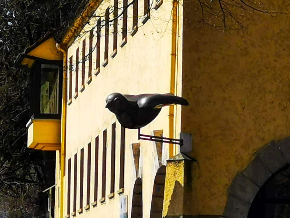 Eine große schwarze Vogelskulptur ist an der gelben Außenwand eines Gebäudes angebracht, über einem gewölbten Eingang mit mehreren Fenstern, die entlang der Wand zu sehen sind - ein auffälliger Ort, der von Besuchern, die die Ulm Card benutzen, oft bemerkt wird.