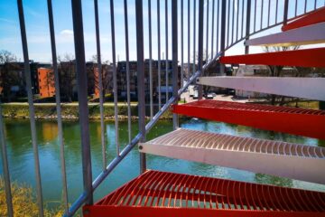 Im Vordergrund ist eine rot-weiße Metalltreppe zu sehen, die auf einen Fluss mit ruhigem Wasser blickt. Auf der anderen Seite des Flusses stehen Ulmer Sehenswürdigkeiten wie moderne Wohnhäuser und blattlose Bäume unter einem blauen Himmel.