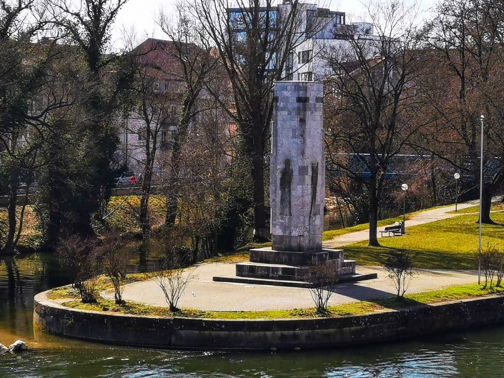 Ein steinernes Denkmal steht in einem kleinen, kreisförmigen Park am Flussufer, umgeben von blattlosen Bäumen und einem Spazierweg, mit Gebäuden im Hintergrund - ein idealer Ort, um mit der Ulm Card auf Entdeckungstour zu gehen.