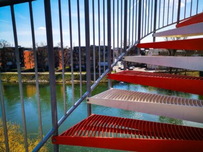 Im Vordergrund ist eine rot-weiße Metalltreppe zu sehen, die auf einen Fluss mit ruhigem Wasser blickt. Auf der anderen Seite des Flusses stehen Ulmer Sehenswürdigkeiten wie moderne Wohnhäuser und blattlose Bäume unter einem blauen Himmel.