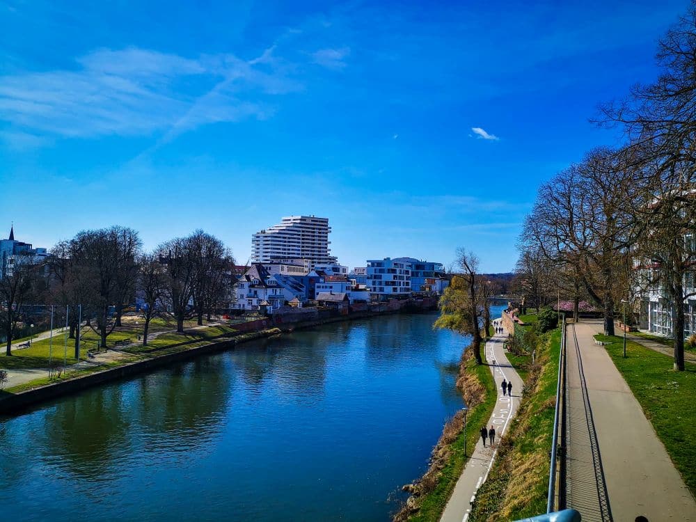 Ein Fluss fließt durch eine Stadt mit modernen weißen Gebäuden und älteren Häusern an den Ufern. Bäume säumen beide Seiten, und mehrere Menschen gehen unter strahlend blauem Himmel auf einem Weg am Wasser entlang, vielleicht mit einer Ulm Card in der Hand.