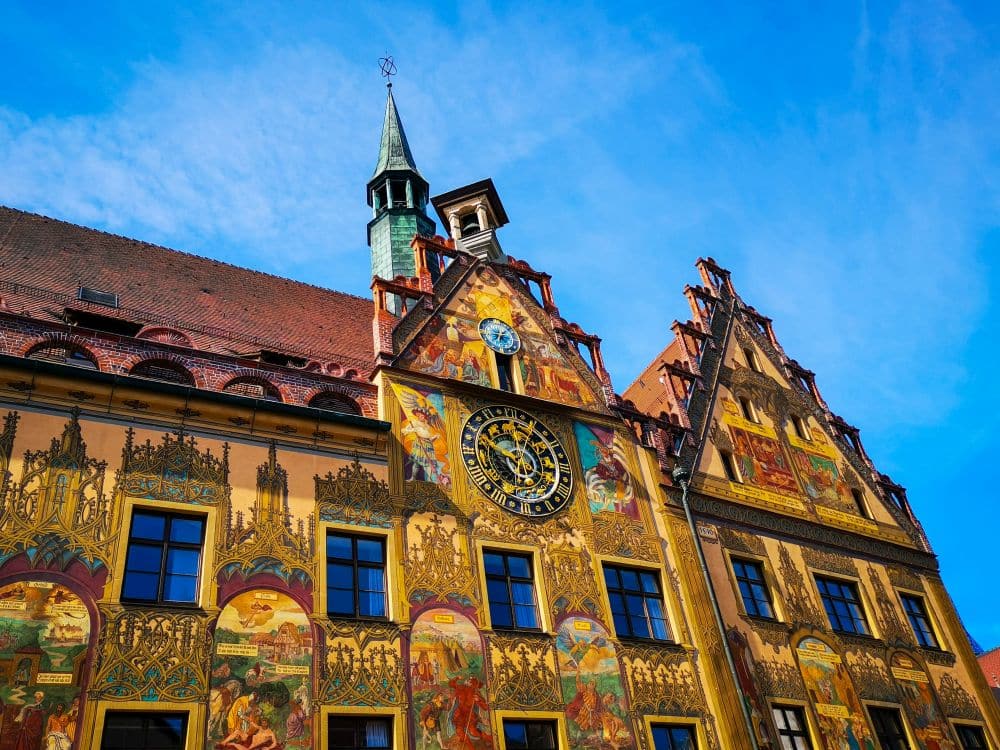 Ein historisches Gebäude mit farbenfrohen Wandmalereien und kunstvollen Details, mit einer großen Uhr an der Fassade, spitzen Dächern und einem kleinen grünen Turm - entdecken Sie es einfach mit der Ulm Card vor einem strahlend blauen Himmel.