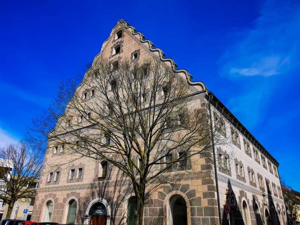Ein großes Steingebäude mit einem steilen, abgestuften Satteldach steht unter einem strahlend blauen Himmel. Laublose Bäume verdecken teilweise die Fassade, und im Erdgeschoss sind gewölbte Türöffnungen zu sehen - ein idealer Anblick für Inhaber der Ulm Card, um ihn zu erkunden.