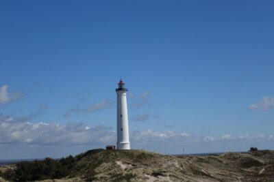 Ein hoher weißer Leuchtturm, Lyngvig Fyr, mit einer roten Spitze steht auf einem grasbewachsenen Hügel unter einem klaren blauen Himmel mit ein paar Wolken. Im Vordergrund sind Sträucher und sandiges Gelände zu sehen.