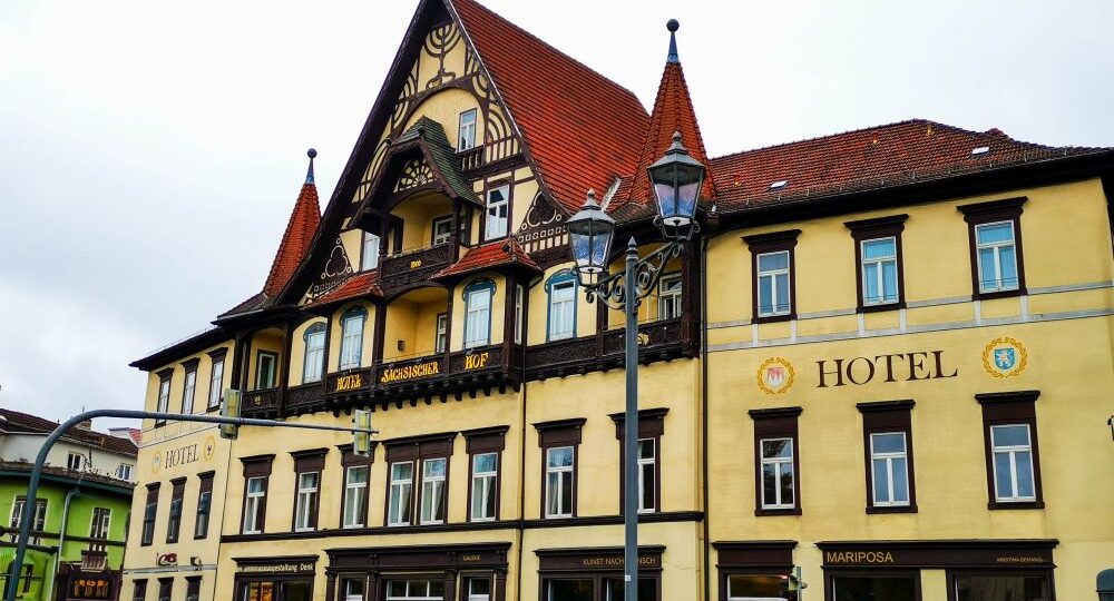 Ein großes, historisches, gelbes Hotel mit braunen Holzakzenten und einem steilen Giebeldach steht an einer Straßenecke unter einem bewölkten Himmel. Dekorative Fenster und Straßenlaternen verleihen ihm Charme - eine einladende Wahl unter den Meininger Hotels.