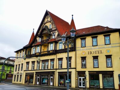 Ein großes, historisches, gelbes Hotel mit braunen Holzakzenten und einem steilen Giebeldach steht an einer Straßenecke unter einem bewölkten Himmel. Dekorative Fenster und Straßenlaternen verleihen ihm Charme - eine einladende Wahl unter den Meininger Hotels.