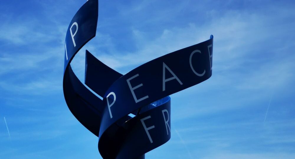 Eine verbogene Metallskulptur vor blauem Himmel, auf deren glatter, dunkler Oberfläche das Wort "PEACE" und Teilbuchstaben zu sehen sind - eine eindrucksvolle Erinnerung an den Kalter Krieg und die historische Bedeutung von Point Alpha.