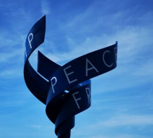 Eine verbogene Metallskulptur vor blauem Himmel, auf deren glatter, dunkler Oberfläche das Wort "PEACE" und Teilbuchstaben zu sehen sind - eine eindrucksvolle Erinnerung an den Kalter Krieg und die historische Bedeutung von Point Alpha.