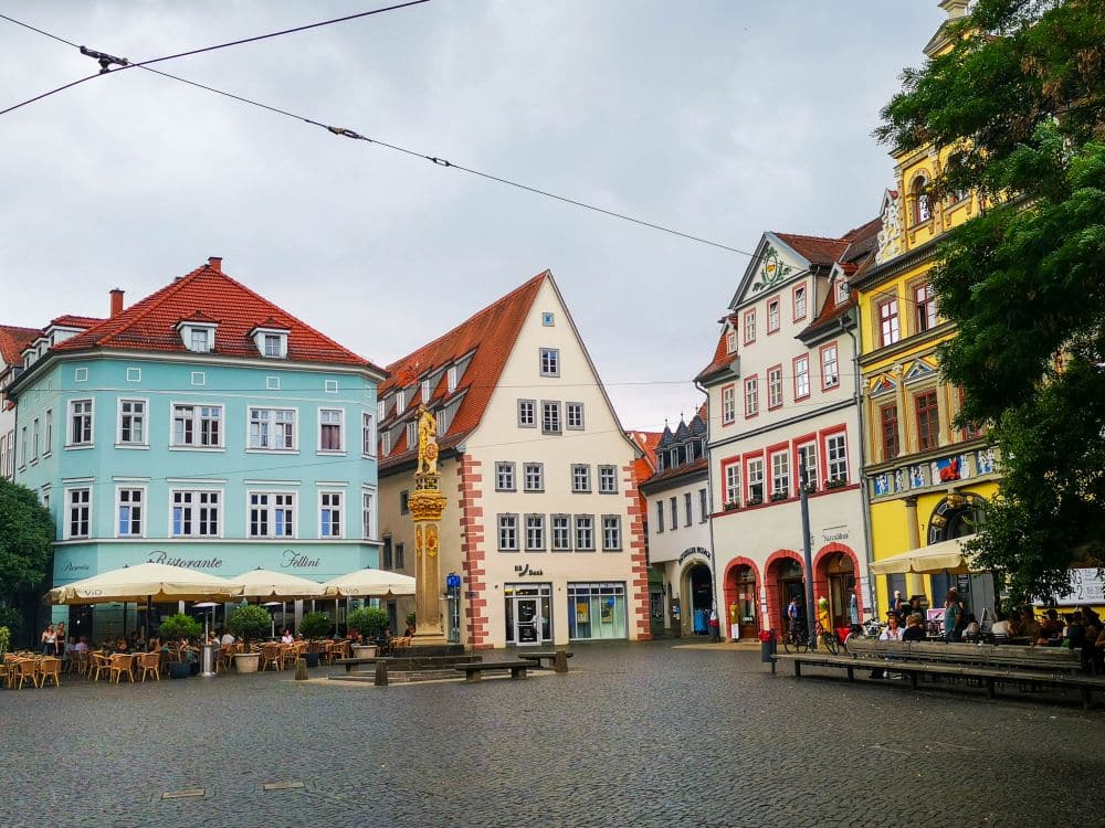 Ein europäischer Stadtplatz in Erfurt mit farbenfrohen historischen Gebäuden, Sitzgelegenheiten in Straßencafés, einer zentralen Statue und Kopfsteinpflaster unter einem bewölkten Himmel. Die Menschen entspannen sich in den Cafés, während Bäume diese charmante Szene einrahmen.