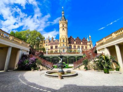 Ein prachtvolles Schloss, das an Schwerin erinnert, mit einem hohen Turm in der Mitte, kunstvoller Architektur und einem Innenhof mit einem runden Brunnen, Topfpflanzen und Treppen vor einem strahlend blauen Himmel mit vereinzelten Wolken.