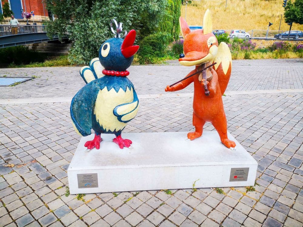 Die farbenfrohen Outdoor-Statuen in Erfurt zeigen einen skurrilen Vogel mit roter Halskette und einen lächelnden orangefarbenen Fuchs, der Geige spielt. Beide stehen auf einem weißen Sockel in einer gepflasterten und begrünten Fläche.