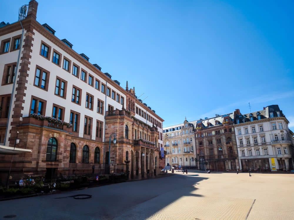 Ein sonniger, leerer Stadtplatz in der hessischen Landeshauptstadt ist umgeben von historischen europäischen Gebäuden mit verzierten Fassaden und großen Fenstern unter einem klaren, blauen Himmel - ideal für Stadtführungen Wiesbaden.