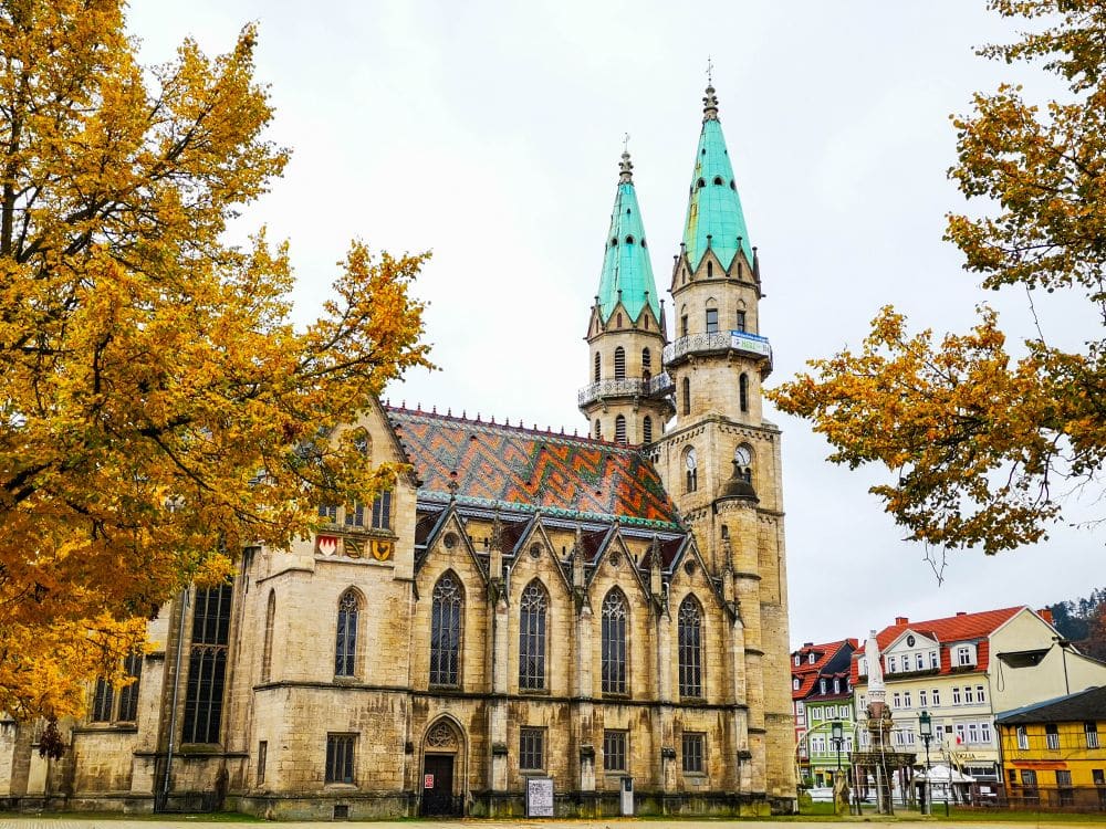 Eine historische Kirche mit bunten, gemusterten Dachziegeln und grünen Zwillingstürmen steht inmitten von Herbstbäumen mit gelben Blättern; in der Nähe bieten traditionelle europäische Häuser charmante Meininger Unterkünfte unter einem wolkenverhangenen Himmel.