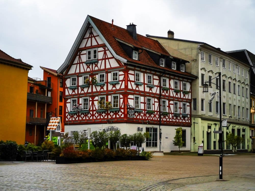 Ein traditionelles Fachwerkhaus mit roten und weißen Mustern steht auf einer Kopfsteinpflasterstraße in Meiningen, umgeben von modernen Gebäuden, Grünanlagen und Sitzgelegenheiten im Freien an einem bewölkten Tag.