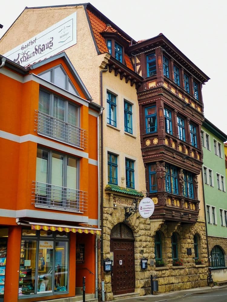 Bunte Gebäude säumen eine Straße in Meiningen; eines davon hat eine obere Fassade aus Fachwerk mit verzierten Holzarbeiten und ein unteres Stockwerk aus Stein. Die angrenzenden Gebäude haben moderne Fenster und leuchtend orange-, grün- und beigefarbene Außenwände. Über den Eingängen hängen Schilder.