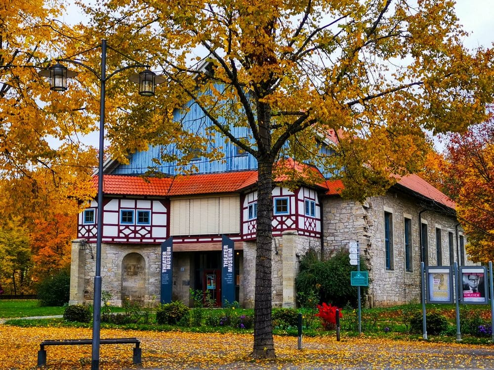 Historisches Gebäude in Meiningen mit Steinmauern, Fachwerk und einem roten Dach, umgeben von bunten Herbstbäumen. Gefallene Blätter bedecken den Boden, in der Nähe stehen Laternenpfähle, eine Bank und Informationsschilder.