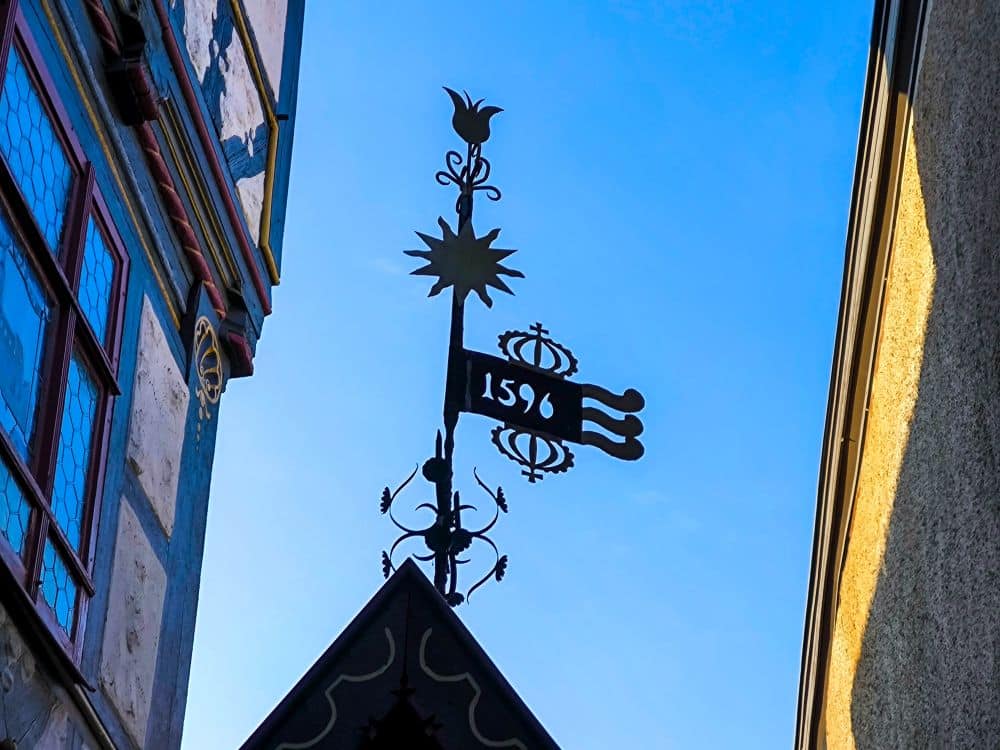 Ein dekoratives schmiedeeisernes Schild mit einer Schlüsselform, der Nummer 1596 und verschnörkelten Details, das sich vor einem blauen Himmel zwischen zwei Gebäuden in Meiningen abhebt. Das eine Gebäude hat bunt bemalte Verkleidungen und Fenster.