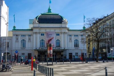 Ein prächtiges weißes Gebäude mit grünem Dach und verzierter Fassade steht in Hamburg, mit Fahnen und einem großen Transparent über dem Eingang, auf dem "Der anarchische Kuss" steht. Fahrräder und Fußgänger füllen den belebten Platz davor.