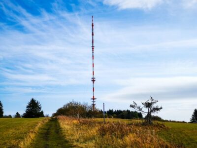 Ein hoher, rot-weißer Funkturm steht auf einer offenen Wiese in der Nähe von Heidelstein, umgeben von Bäumen und einem blauen Himmel mit Wolkenfetzen. Dünne Kabel stützen den Turm, und ein Feldweg führt zu ihm hin.