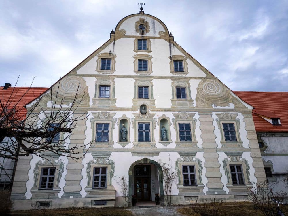 Ein historisches Barockgebäude mit einer dekorativen weißen Fassade, geometrischen und spiralförmigen Mustern, einem gewölbten Eingang und mehreren Statuen in Nischen. Ein steiles rotes Dach unter einem wolkenverhangenen Himmel - kahle Bäume stehen vor dem Kloster Benediktbeuern, dahinter liegen die Berge.