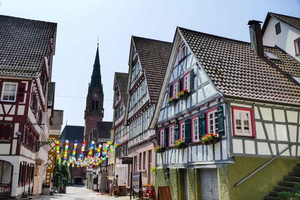 Eine charmante Straße in Calw mit traditionellen Fachwerkhäusern, Blumenkästen an den Fenstern und bunten Hängedekorationen. Im Hintergrund erhebt sich eine Kirche mit einem hohen Kirchturm unter einem klaren Himmel.