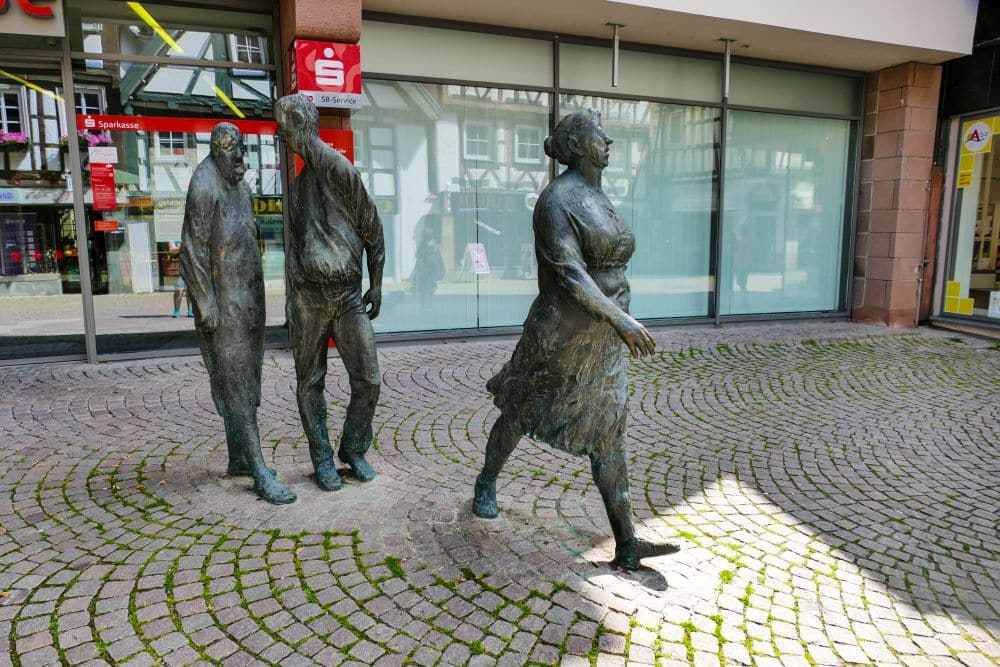 Drei lebensgroße Bronzestatuen stehen auf einer Kopfsteinpflasterstraße in Calw; zwei Männer scheinen eine Frau zu beobachten, die zwischen modernen Glasfassaden und einem Bankschild vor ihnen hergeht.