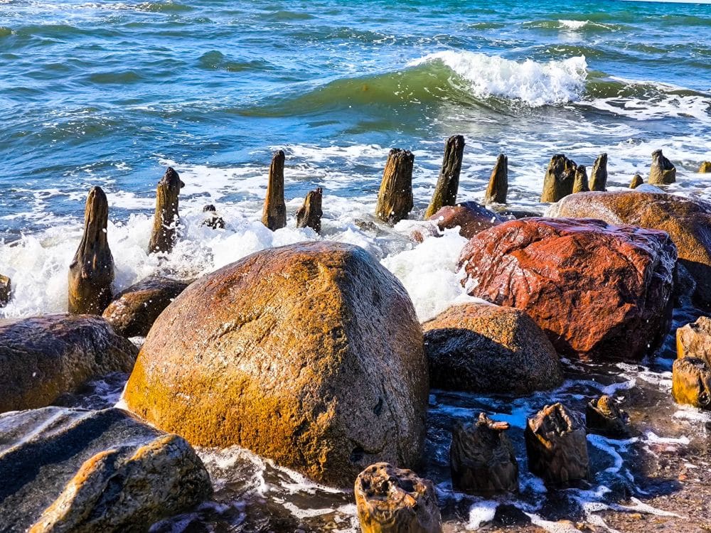 Große Felsen und Holzpfähle säumen das Kühlungsborner Ufer, an dem die Wellen anschlagen, während im Hintergrund das blaugrüne Meerwasser unter einem hellen, sonnigen Himmel zu sehen ist.