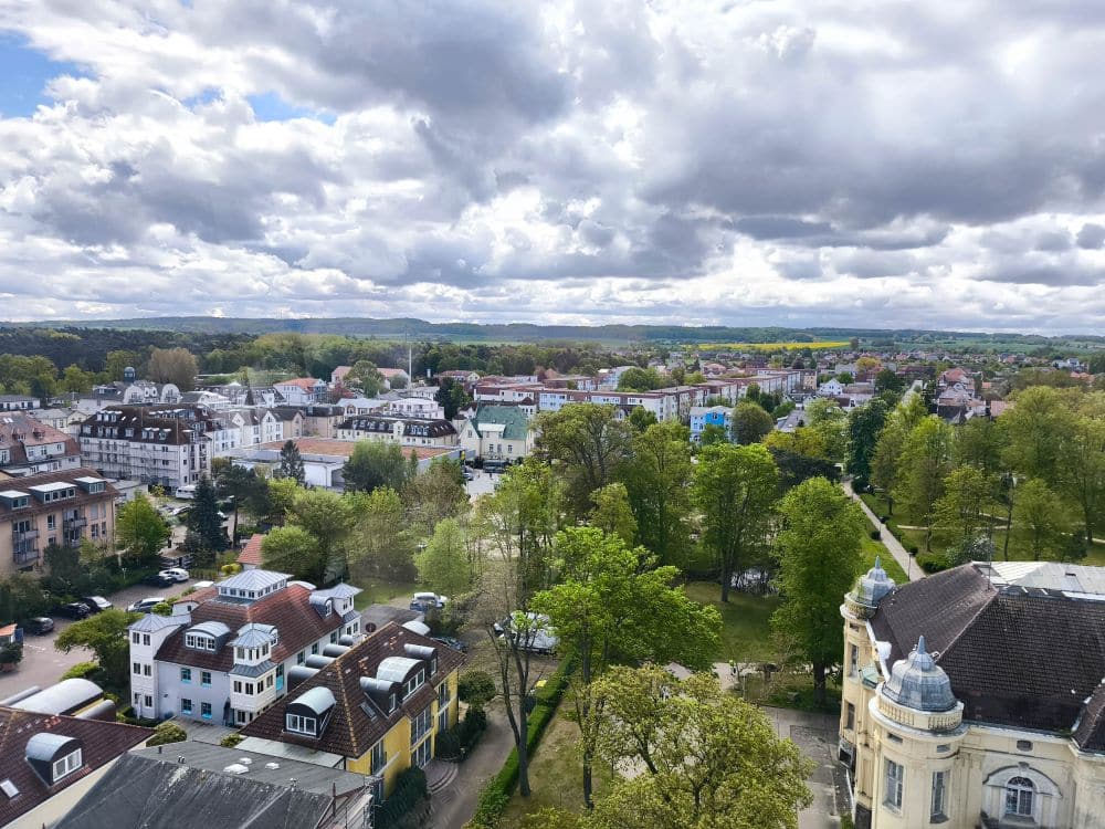Luftaufnahme von Kühlungsborn mit Häusern, Bäumen und einem Park bei teilweise bewölktem Himmel, eingerahmt von weit entfernten grünen Feldern und Hügeln am Horizont.