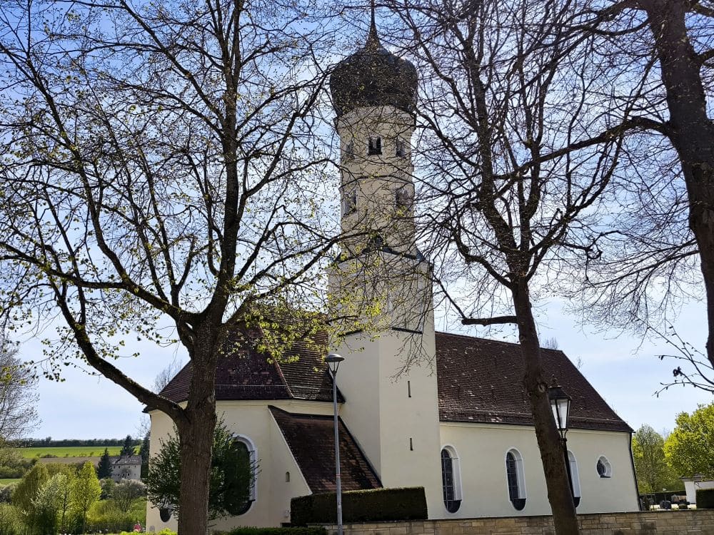 Eine weiße Kirche mit einem hohen, zwiebelförmigen Kirchturm steht an einem sonnigen Tag in der Klosterstadt Neresheim hinter blattlosen Bäumen. Das braune Dach und die gewölbten Fenster verleihen ihr einen besonderen Charme, während die grünen Felder und der blaue Himmel das ruhige Bild vervollständigen.