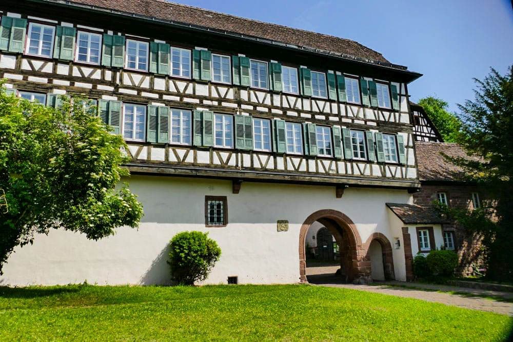 Ein großes Fachwerkhaus mit grünen Fensterläden und vielen Fenstern steht hinter einem bogenförmigen Steintor in Hirsau, umgeben von grünem Gras, Bäumen und Büschen an einem sonnigen Tag.